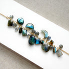 A gold chain necklace featuring natural labradorite gemstones in various shapes and sizes, displayed on a white rectangular card over a textured gray fabric background.