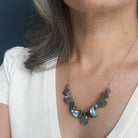 A close-up of a person wearing a gold chain necklace with faceted gray-blue labradorite stones, styled with a white top against a dark blue background.