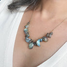 A close-up of a gold chain necklace with irregular, faceted blue-gray labradorite stones worn by a model in a white top.