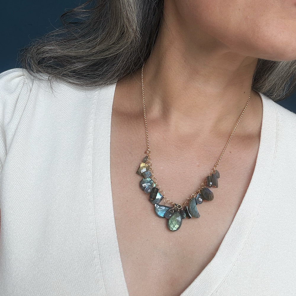 A close-up of a woman wearing a gold chain necklace with irregular, shimmering gray-green stones, paired with a white top.