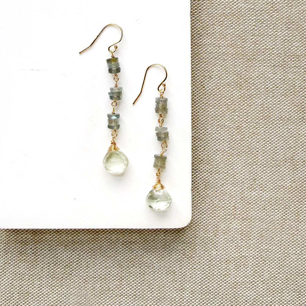 A pair of handmade dangle earrings with greenish-gray stone beads and clear teardrop crystals, displayed on a white rectangular block over a beige woven fabric background.