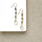A pair of handmade dangle earrings with greenish-gray stone beads and clear teardrop crystals, displayed on a white rectangular block over a beige woven fabric background.