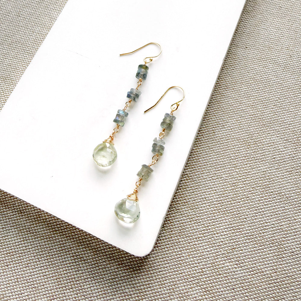 Dangle earrings with small blue-green faceted beads and clear teardrop-shaped crystals on gold hooks, displayed on a white card over a beige woven fabric background.