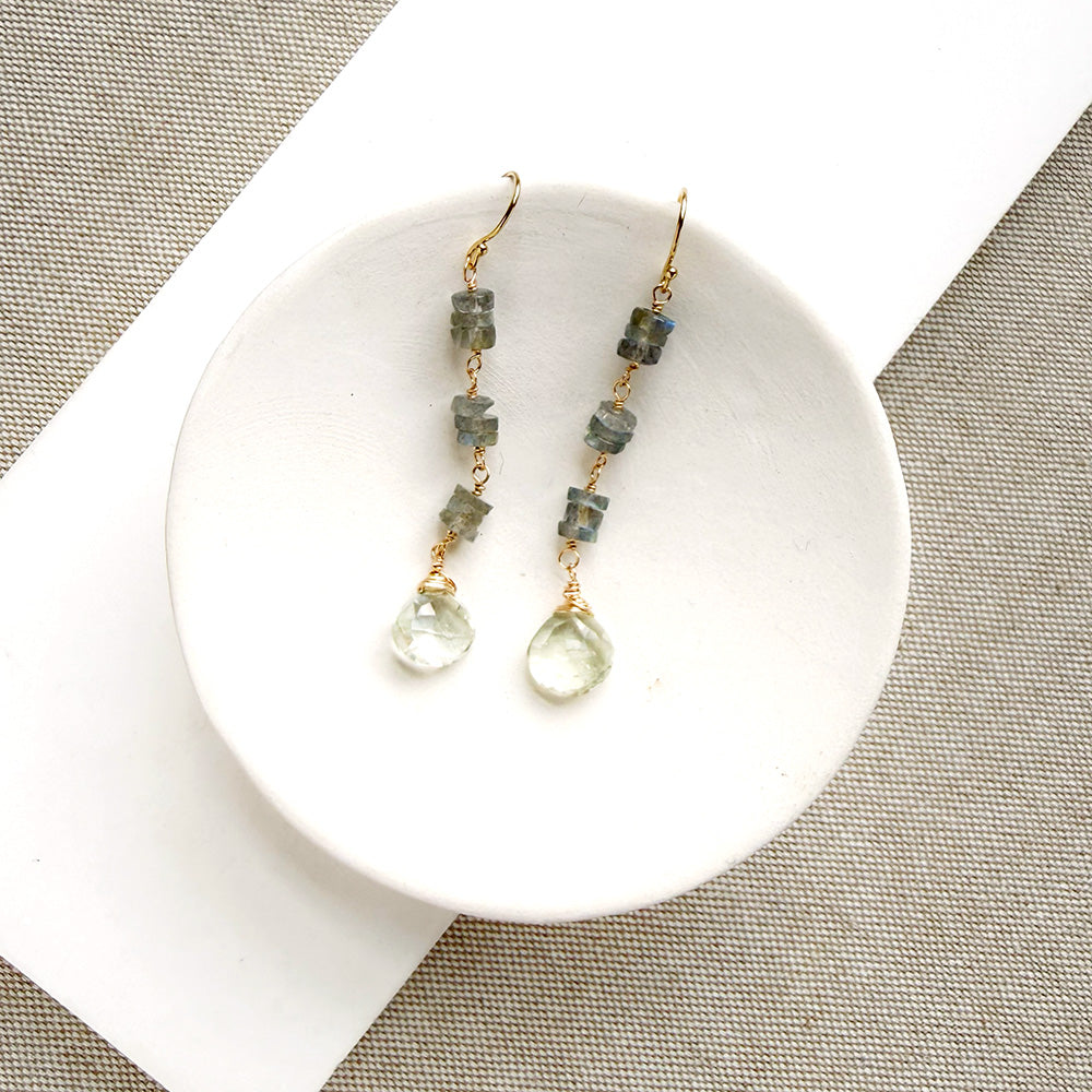 A pair of gold-tone dangle earrings with small gray-green stone beads and clear faceted teardrop drops, displayed on a white ceramic dish over a textured beige fabric background.