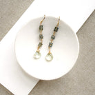 A pair of gold-tone dangle earrings with small gray-green stone beads and clear faceted teardrop drops, displayed on a white ceramic dish over a textured beige fabric background.