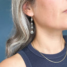 A close-up of a woman wearing a delicate gold drop earring with small clear faceted beads and a larger clear teardrop charm. The earring is shown on her ear, with her gray hair and navy blue top visible against a soft blue-gray background.
