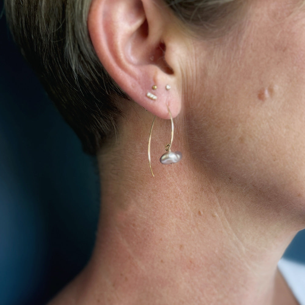 A close-up of a person's ear wearing a delicate gold huggie hoop earring with a small silver baroque pearl dangle, alongside other ear piercings.