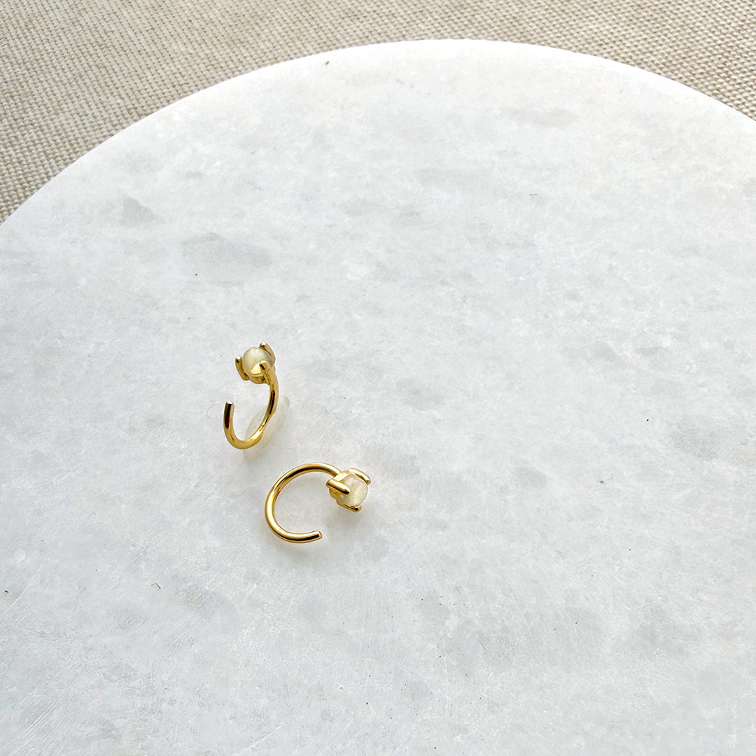 Two tiny gold reverse huggie style earring with opal stones on a round marble surface with a beige fabric background