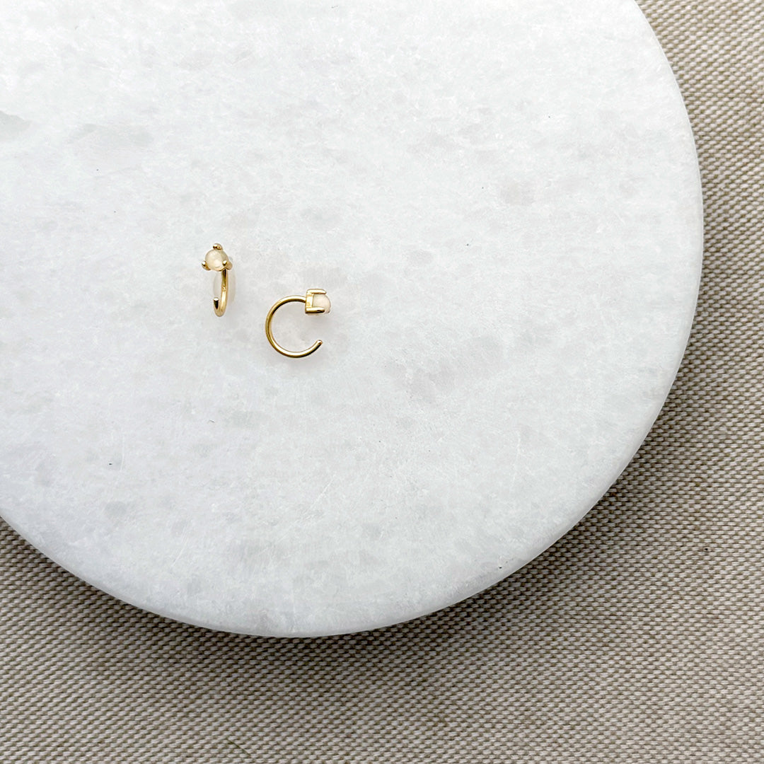 Two tiny gold reverse huggie style earring with opal stones on a round marble surface with a beige fabric background