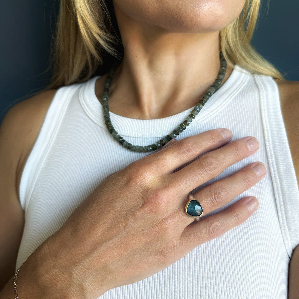 A close-up lifestyle shot of a woman wearing a dark green beaded necklace and a london blue topaz ring with a teardrop-shaped stone on her finger, paired with a white ribbed tank top.