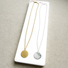 Two round pendant necklaces displayed on a white rectangular card against a neutral beige fabric background. One necklace is gold with a hammered disc pendant, and the other is silver with a hammered disc pendant.