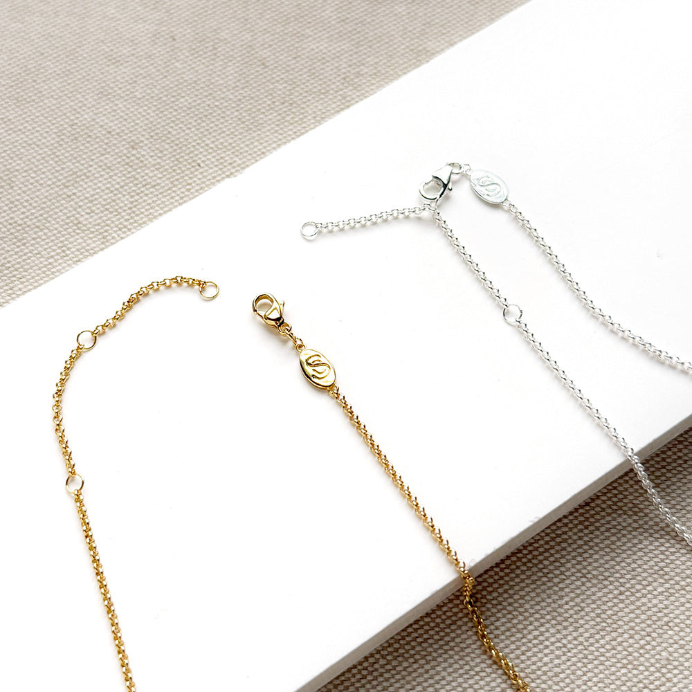 Close-up of two delicate chain necklaces, one gold-toned and one silver-toned, laid on a white rectangular surface with a beige textured fabric background. Each chain includes a lobster clasp, small circular links, and a round branded tag.
