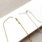 Close-up of two delicate chain necklaces, one gold-toned and one silver-toned, laid on a white rectangular surface with a beige textured fabric background. Each chain includes a lobster clasp, small circular links, and a round branded tag.