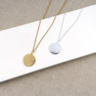 Two minimalist hammered round pendant necklaces displayed on a white rectangular block over a beige textured fabric background. One necklace has a gold chain and a gold pendant, and the other has a silver chain and a silver pendant.