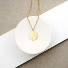 A gold chain necklace with a textured, hammered circular pendant, displayed on a white ceramic dish over a neutral beige and white background.