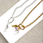 Two delicate chain necklaces displayed on a white rectangular card against a beige textured fabric background. One necklace is silver with a fish charm and a small clear crystal pendant.