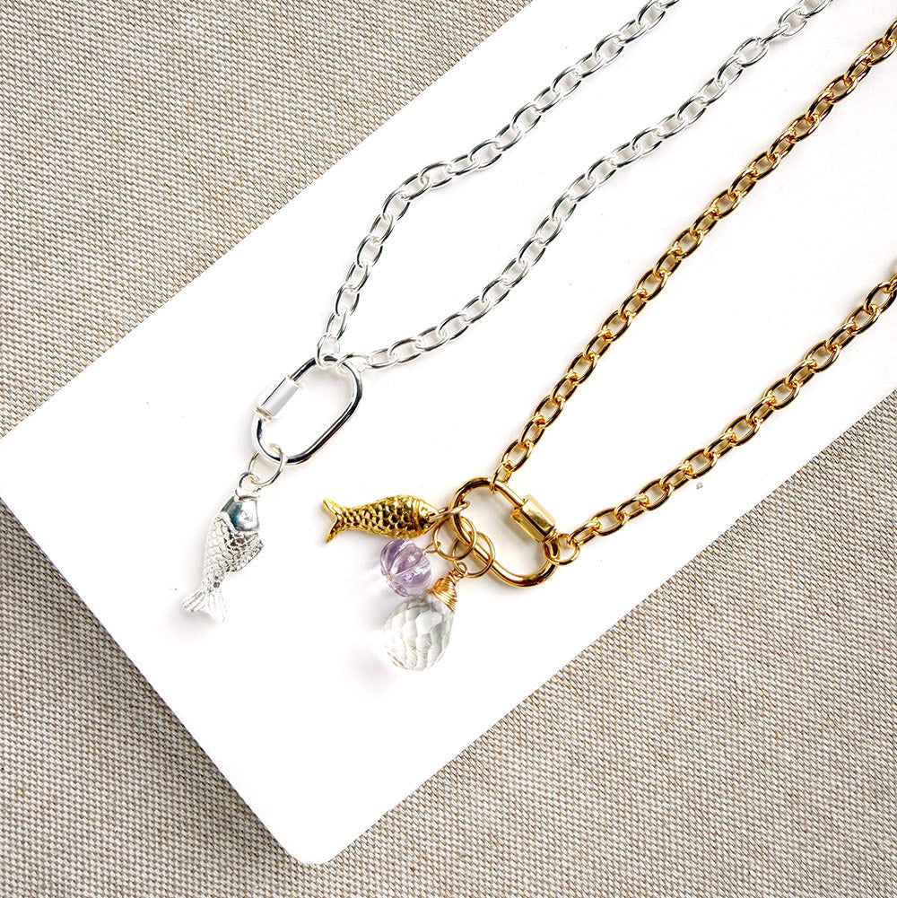 Two chain necklaces displayed on a white rectangular card against a beige textured fabric background. One necklace is silver with a fish charm and a small clear crystal pendant.