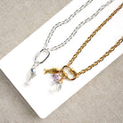 Two chain necklaces displayed on a white rectangular card against a beige textured fabric background. One necklace is silver with a fish charm and a small clear crystal pendant.