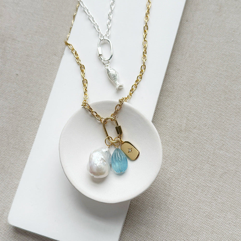A gold chain necklace with a fish-shaped charm and three dangling charms (a white pearl, a faceted light blue stone, and a small gold tag) displayed on a white dish over a neutral beige fabric background.