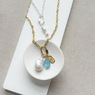 A gold chain necklace with a fish-shaped charm and three dangling charms (a white pearl, a faceted light blue stone, and a small gold tag) displayed on a white dish over a neutral beige fabric background.