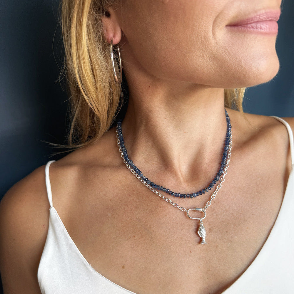 A close-up of a woman wearing layered necklaces with a silver chain and a blue kyanite gemstones featuring a small silver pendant, paired with a white camisole top.