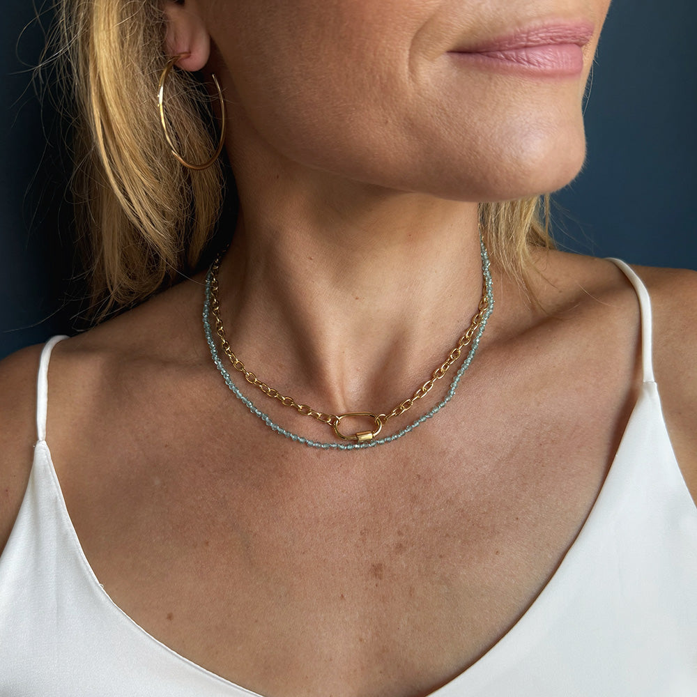 A close-up of a woman wearing a double-layered necklace. The necklace features one layer of gold chain with a small gold oval charm and the other layer of textured light blue beads.