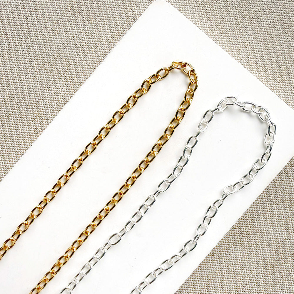 Two metal chains, one gold-toned and one silver-toned, laid diagonally on a white rectangular card over a beige textured fabric background.