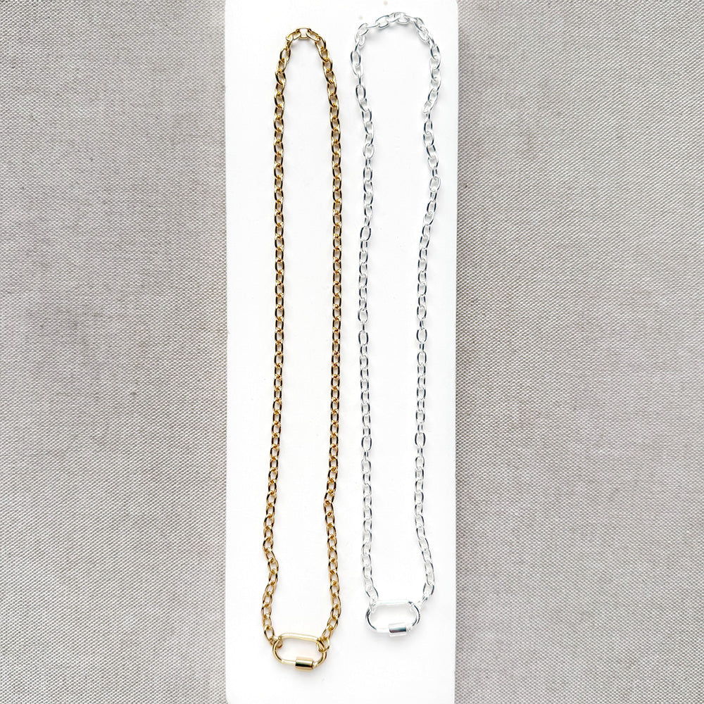 Two chain necklaces, one gold and one silver, displayed on a white rectangular card against a textured gray fabric background.