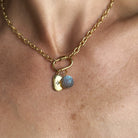 A close-up of a gold chain necklace worn on a person's neck, featuring a small gold tag charm with a star and a round carved labradorite charm.