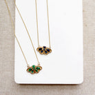 Two delicate gold chain necklaces with butterfly-shaped pendants, one featuring green stones and the other blue stones, displayed on a white card over a beige textured fabric background.