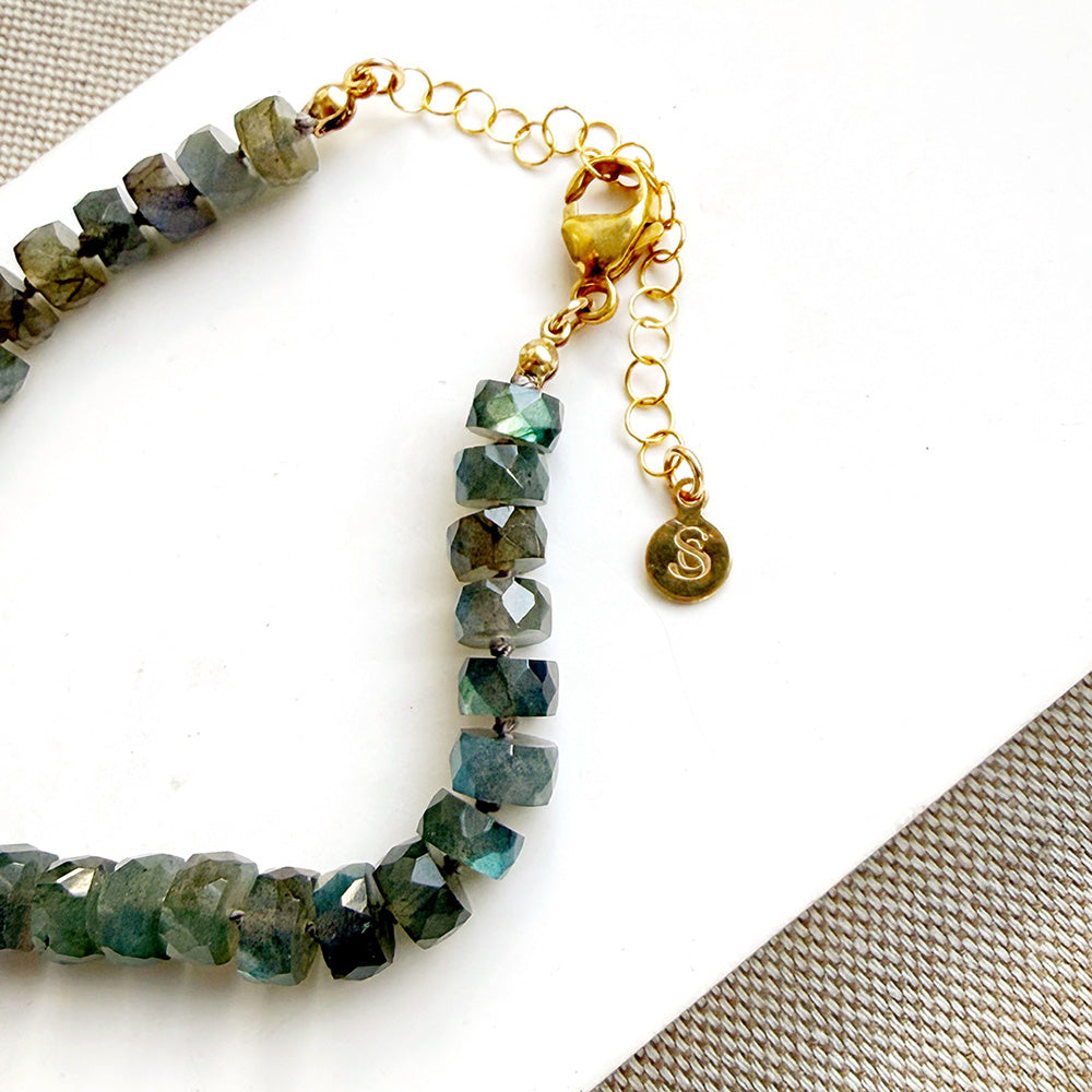 Faceted green and blue labradorite beads on a gold chain bracelet with a lobster clasp and a small engraved gold charm, displayed on a white surface.