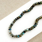 Faceted round gemstone beads with a blue-green iridescent finish, arranged in a curved strand on a white surface with a beige textured fabric background.