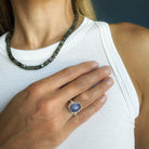 A close-up of a person wearing a silver ring with a tanzanite cabochon stone, paired with a dark green beaded necklace and a white ribbed tank top.