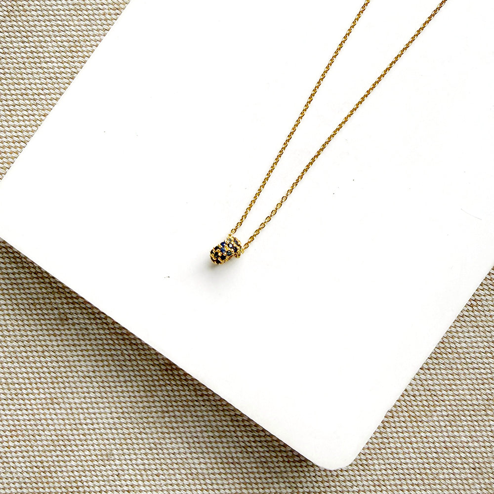 A delicate gold chain necklace with a pave sapphire charm, displayed on a white square card over a beige woven fabric background.