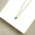 A delicate gold chain necklace with a pave sapphire charm, displayed on a white square card over a beige woven fabric background.