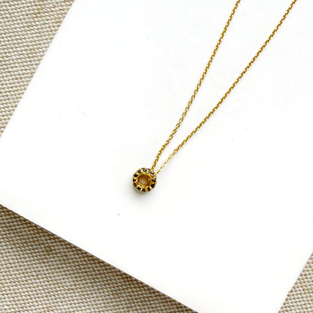 A delicate gold chain necklace with a pave sapphire charm, displayed on a white square card over a beige woven fabric background.