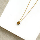 A delicate gold chain necklace with a pave sapphire charm, displayed on a white square card over a beige woven fabric background.