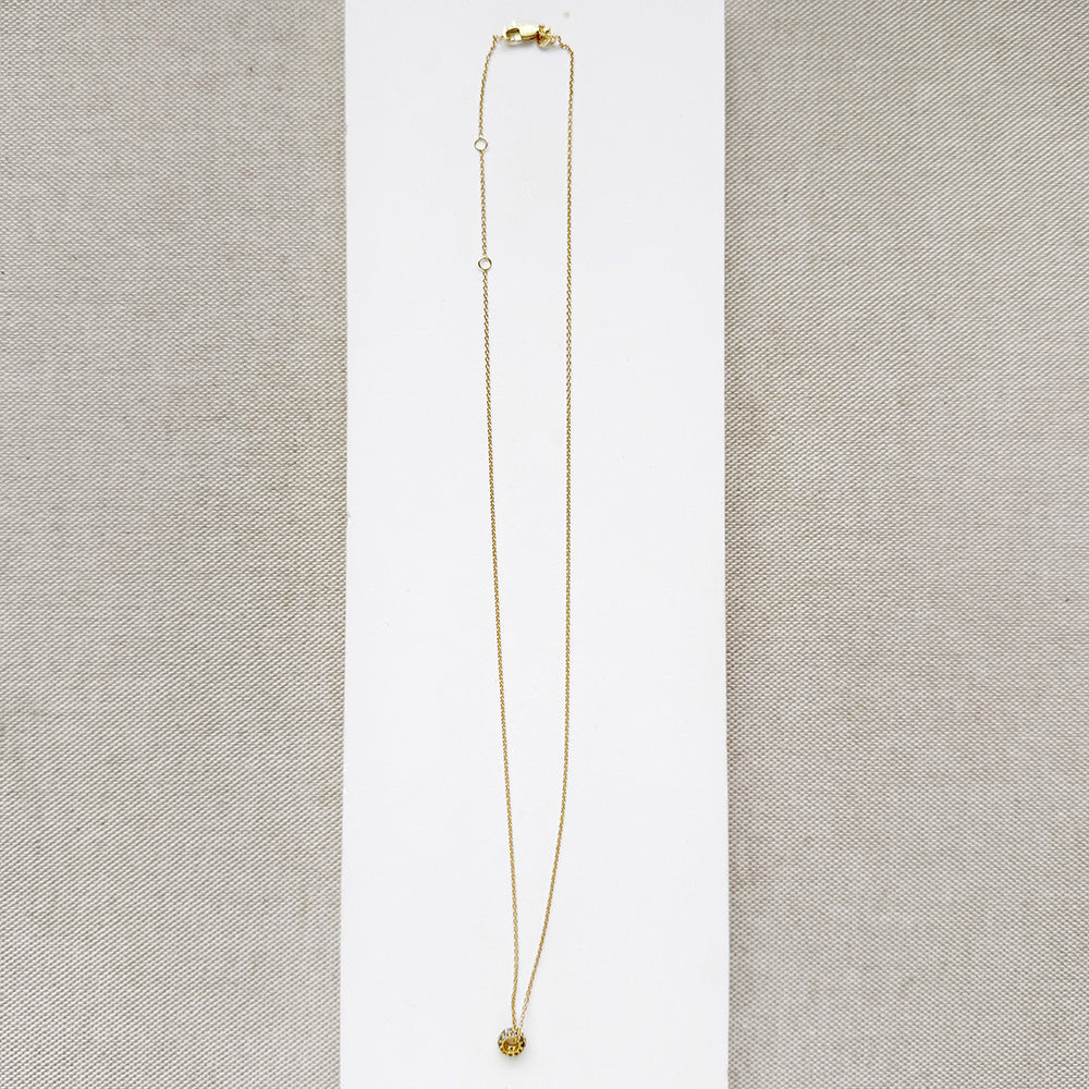 A delicate gold chain necklace with a pave sapphire charm, displayed on a white square card over a beige woven fabric background.