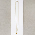 A delicate gold chain necklace with a pave sapphire charm, displayed on a white square card over a beige woven fabric background.