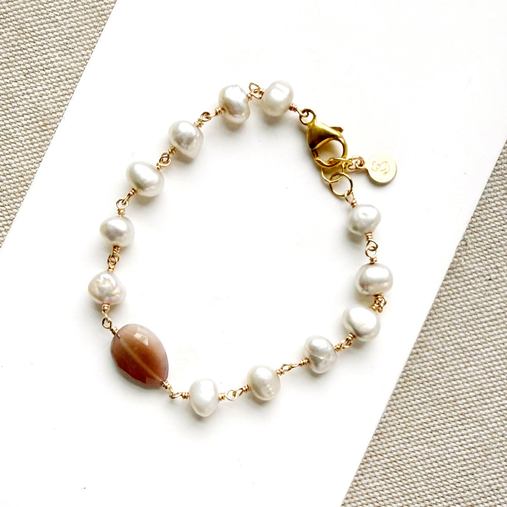 A gold bracelet featuring white pearls and a polished coffee moonstone bead, displayed on a white textured surface.