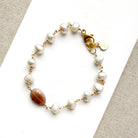 A gold bracelet featuring white pearls and a polished coffee moonstone bead, displayed on a white textured surface.