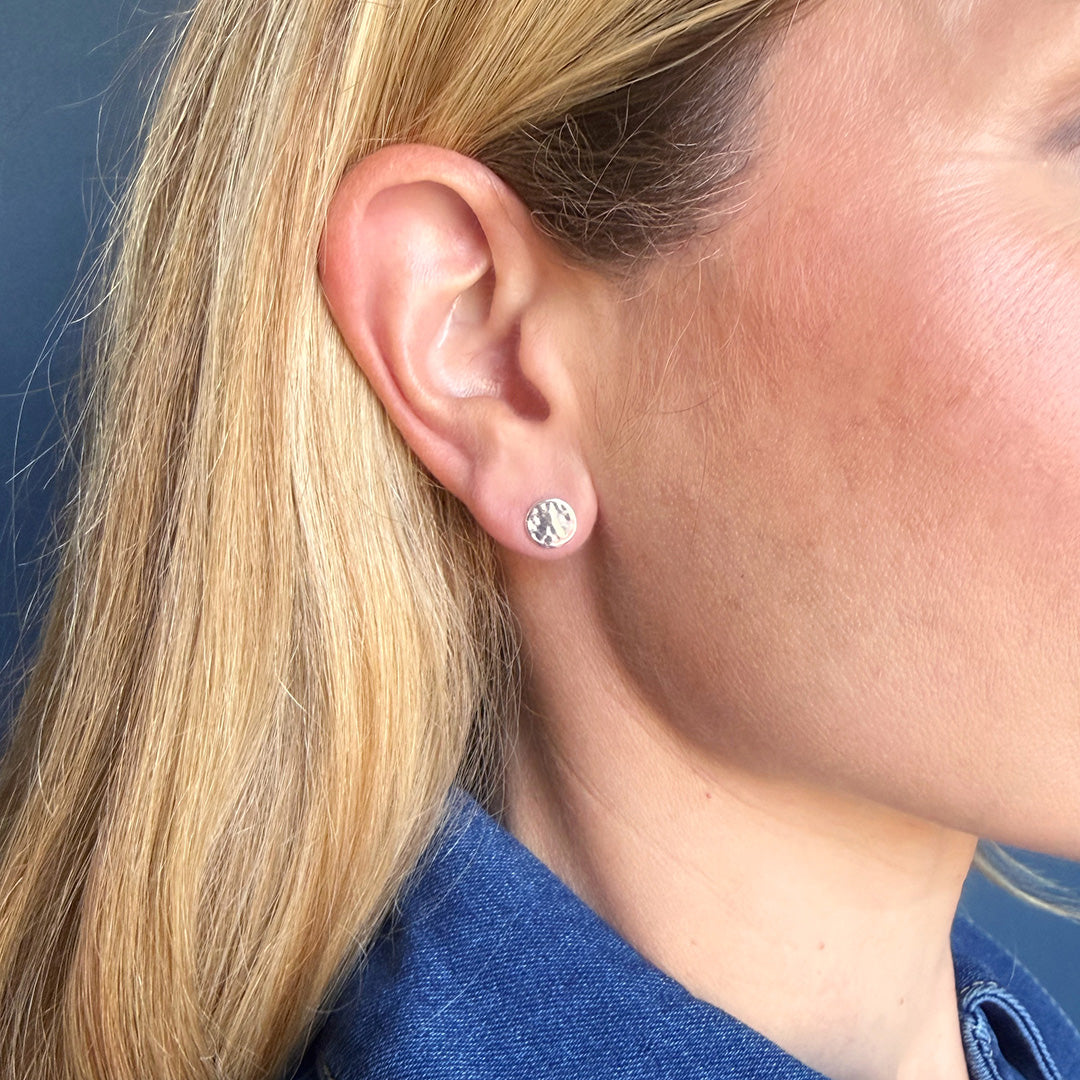 Close-up of a person wearing a silver hammered disc earring against a blue background.