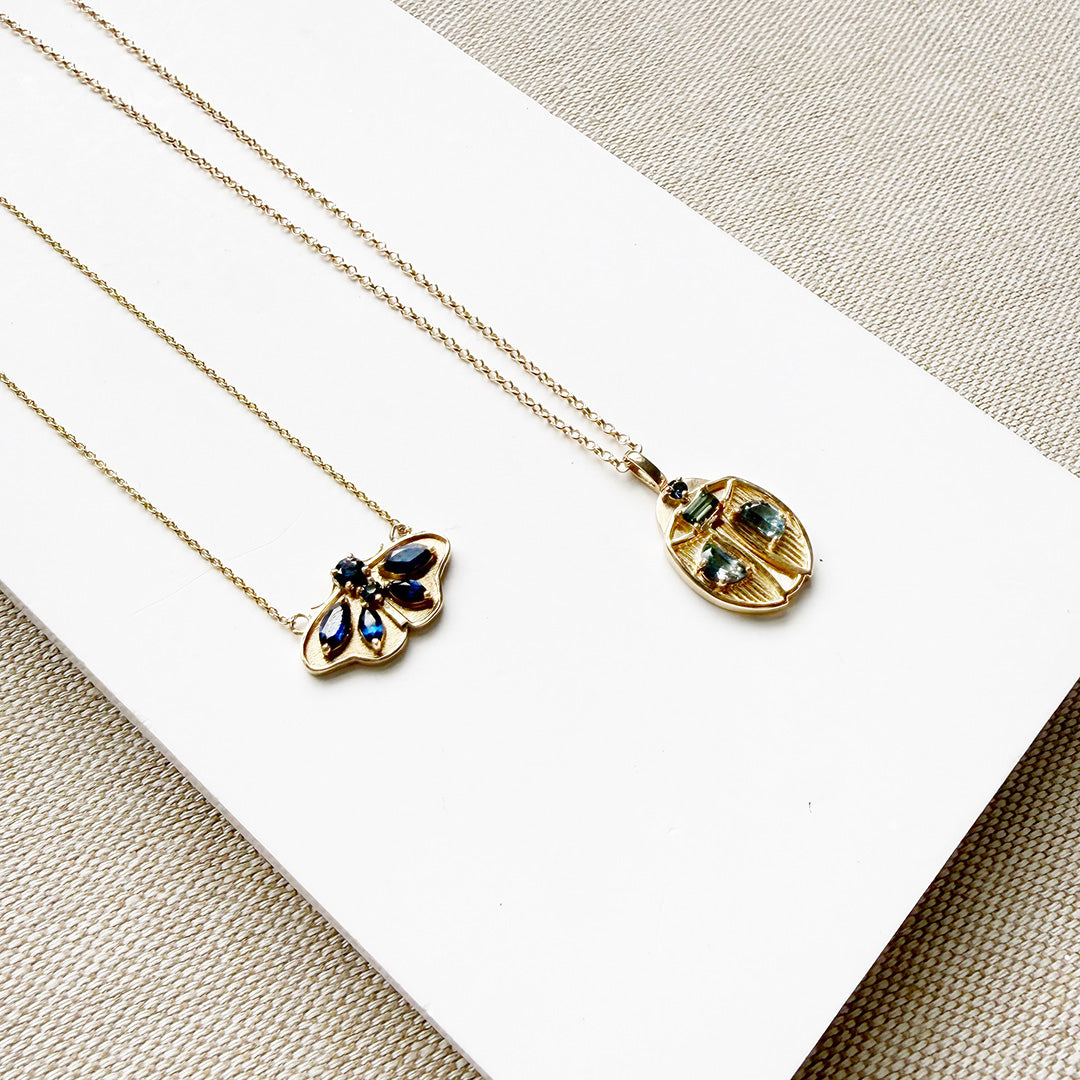 Two gold necklaces with butterfly and beetle pendants on a white surface