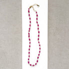 A necklace featuring pink sapphires linked by gold-toned metal, displayed on a white rectangular card over a textured beige fabric background.