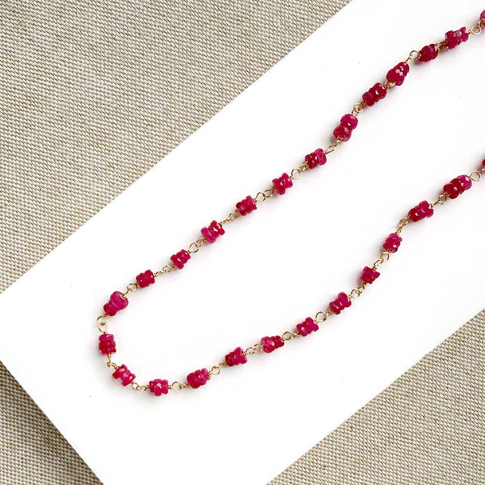 A necklace featuring pink sapphires linked by gold-toned metal, displayed on a white rectangular card over a textured beige fabric background.