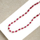 A necklace featuring pink sapphires linked by gold-toned metal, displayed on a white rectangular card over a textured beige fabric background.
