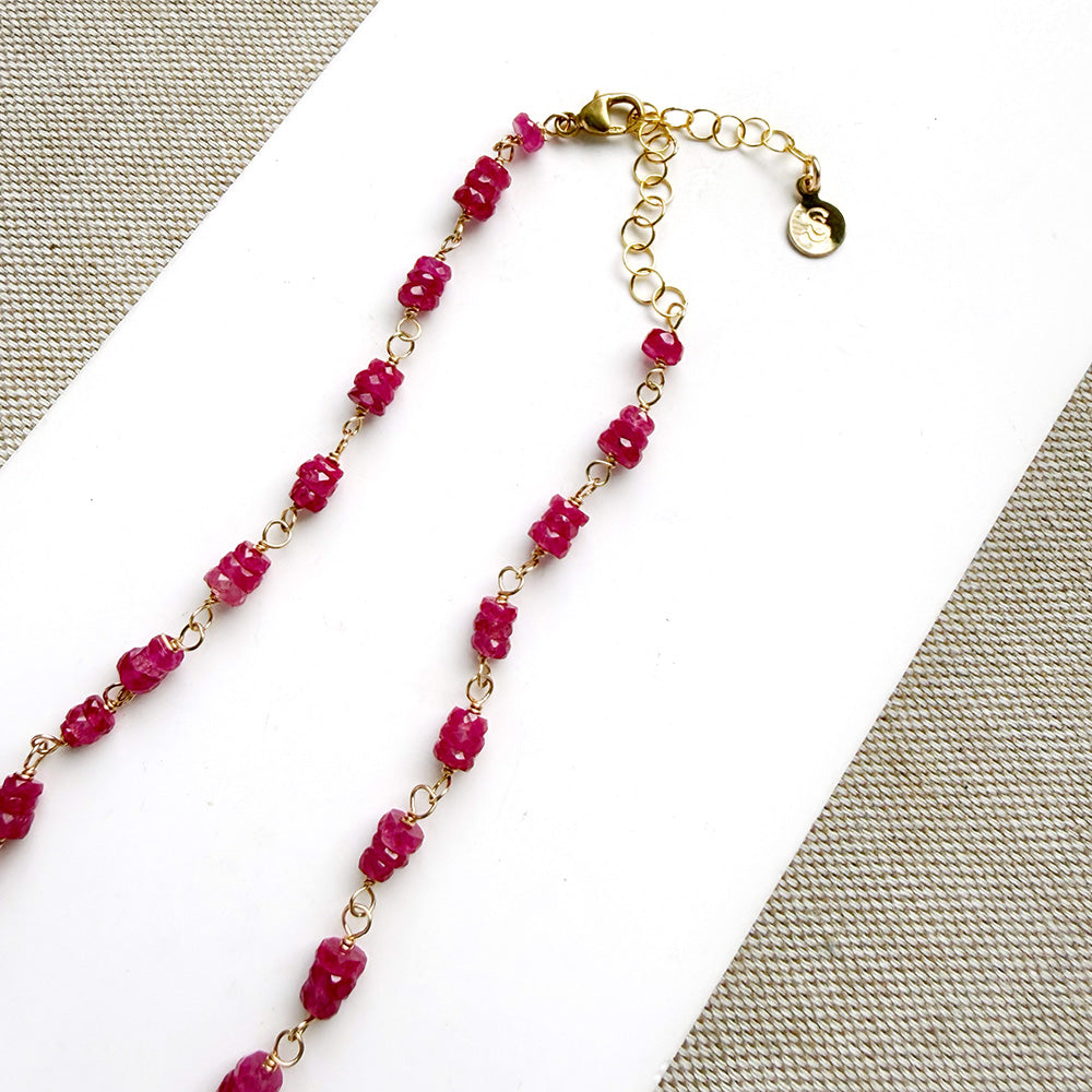 A necklace featuring pink sapphires linked by gold-toned metal, displayed on a white rectangular card over a textured beige fabric background.