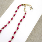 A necklace featuring pink sapphires linked by gold-toned metal, displayed on a white rectangular card over a textured beige fabric background.