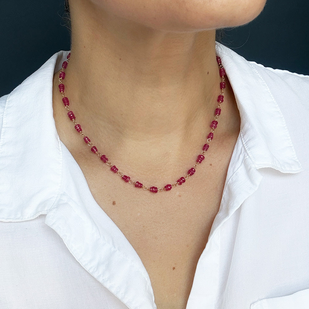 A close-up of a person wearing a pink sapphire necklace, styled with a white collared shirt against a dark background.