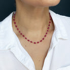 A close-up of a person wearing a pink sapphire necklace, styled with a white collared shirt against a dark background.